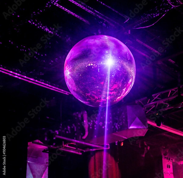 Fototapeta A shimmering disco ball illuminated by purple party lights hangs from the ceiling of a nightclub, creating a vibrant and festive atmosphere