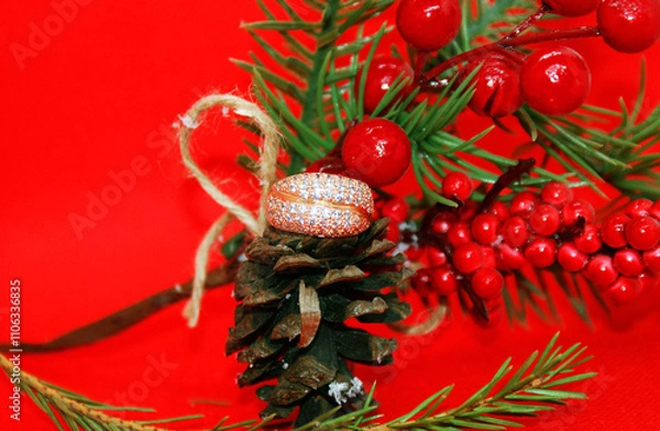 Fototapeta Wedding ring and a spruce branch with a pine cone on a red background, holiday, Christmas.