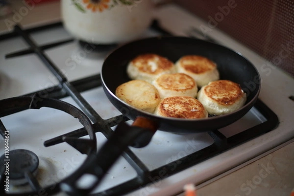 Obraz Golden-brown cheese pancakes frying in a black pan on a gas stove. A retro-styled teapot is visible in the background, creating a cozy and homely atmosphere
