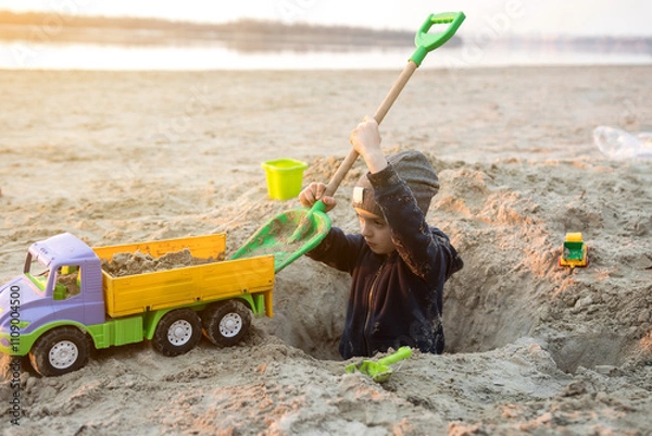 Fototapeta A child playing on the beach with a toy truck and shovel, digging a hole in the sand. The scene captures the joy and creativity of outdoor playtime by the water.