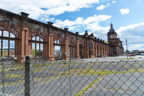 Fototapeta A vacant parking lot fronts a decayed red-brick facade with arched windows, framed by a wire fence. The building’s timeless architecture contrasts with its weathered state under a bright blue sky.