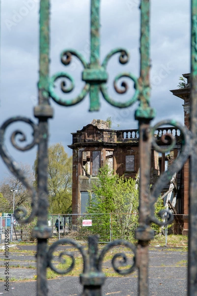 Fototapeta An ornate green metal gate frames a decaying red-brick building with shattered windows, overgrown foliage, and a “Keep Out” sign. The scene conveys abandonment against a cloudy sky.