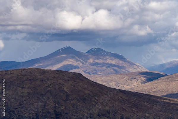 Fototapeta This image features rugged, sunlit mountain peaks with patches of snow, surrounded by shadowed hills under a dramatic sky of thick, brooding clouds, creating a striking and moody scene.