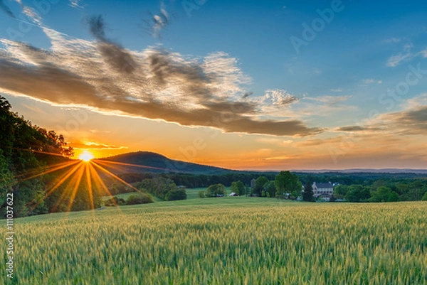 Fototapeta Idealny wiosenny dzień kończy się, gdy słońce zachodzi i rozpyla ciepłe, złote światło na piękną farmę z wyrastającymi polami, kamiennym domem i górskimi grzbietami w tle.