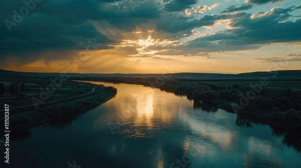 Fototapeta Serene Sunset Over Calm River Surrounded by Lush Greenery and Open Fields with Dramatic Cloud Patterns Illuminated by Golden Rays of Sunlight