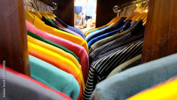 Fototapeta A colorful display of clothing items hanging on wooden hangers in a clothing store. The shirts are arranged in rows, with each row featuring a different color