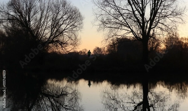 Obraz A man is running in nature just before sunrise. A wooded area with water. You can see the reflection of the man and the trees in the water. (location: Meinerswijk, Arnhem, the Netherlands)