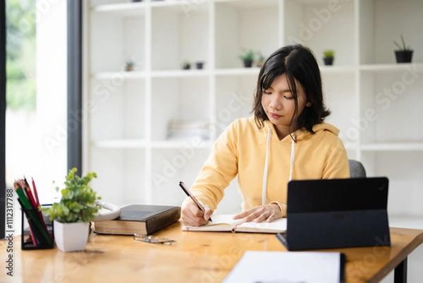 Fototapeta education, online, studying, E learning, young Asian woman having video call on her computer at home. Girl reading book preparing exam test having online consultation, education and learning concept.