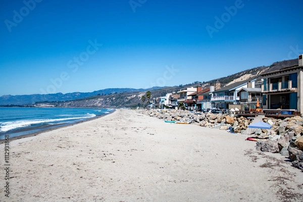 Obraz Beachfront homes along the California coast with a sandy shoreline and scenic mountain backdrop under clear skies.
