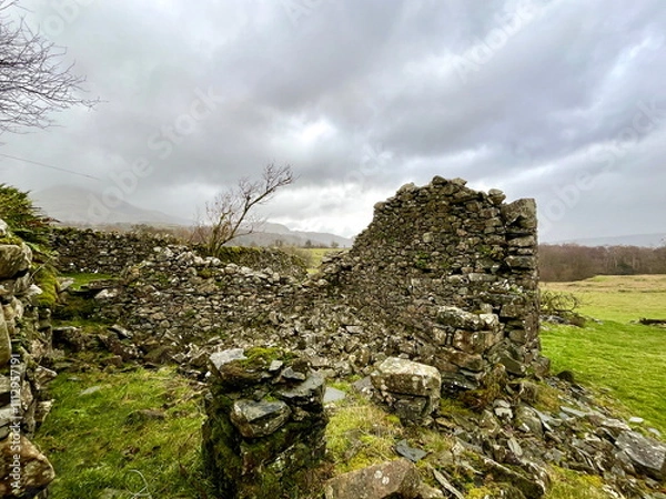 Fototapeta The weathered ruins of a stone building on a grassy hillside, set against a dramatic cloudy sky, showcasing historical decay.