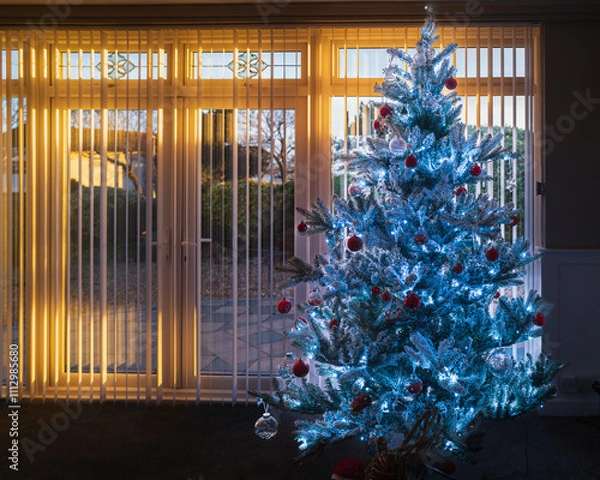 Fototapeta Pretty frosted artificial Christmas tree with fairy lights during a golden sunset  seen through a patio window with vertical slat blinds.