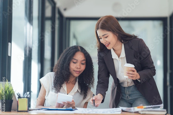 Fototapeta Collaborative Spirit: Two diverse businesswomen, one with a coffee cup in hand, engage in a productive discussion over blueprints.