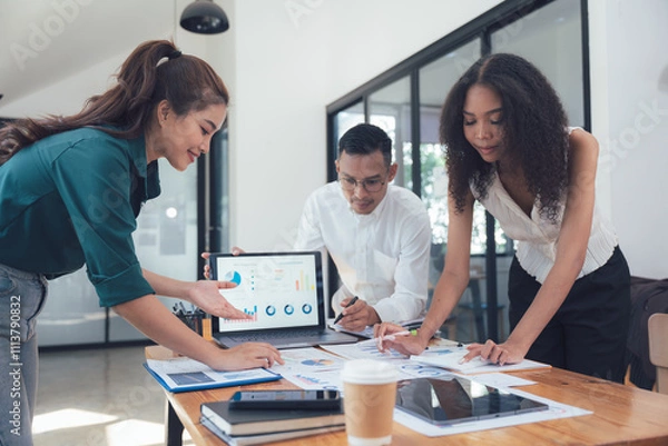 Obraz Collaborative Strategy: A diverse team of professionals huddle around a laptop, analyzing data and charts in a modern office setting. They are engaged in a collaborative discussion.