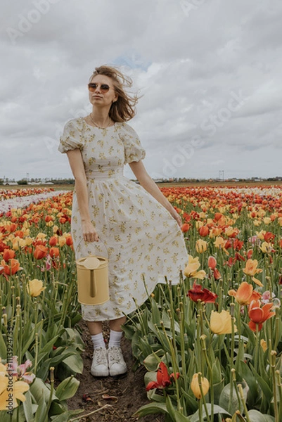 Fototapeta A girl in a yellow dress and apron with a water can in a red and green tulip flower field on a gloomy rainy cloudy sky background happily running around. 