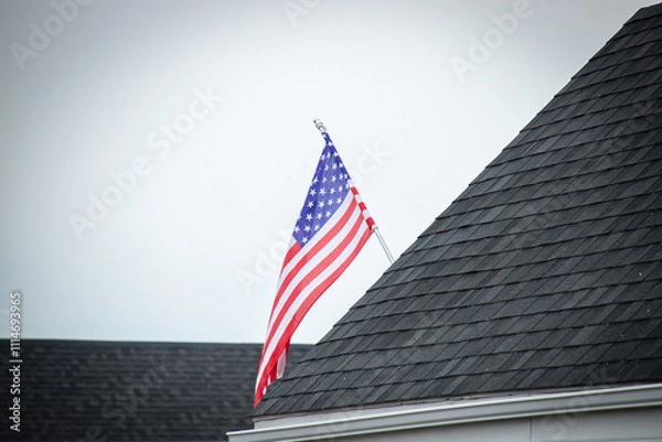 Fototapeta An American flag mounted on a rooftop with dark shingles, set against a cloudy sky, symbolizing patriotism and pride.