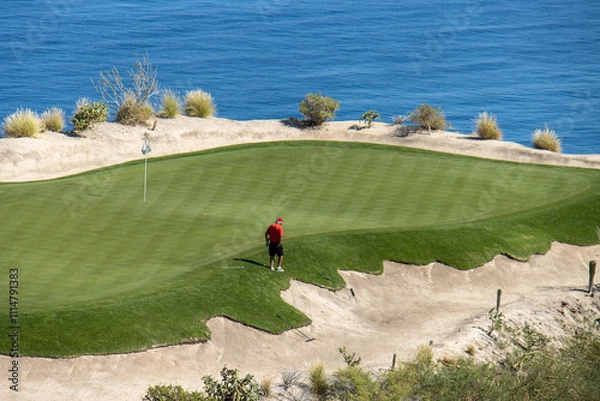 Fototapeta overview of man in red shirt on the edge of putting green with water in  the background