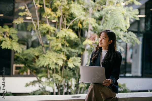 Fototapeta Young businesswoman sits with her laptop in a modern office space adorned with lush greenery, looking thoughtfully towards upcoming projects and opportunities