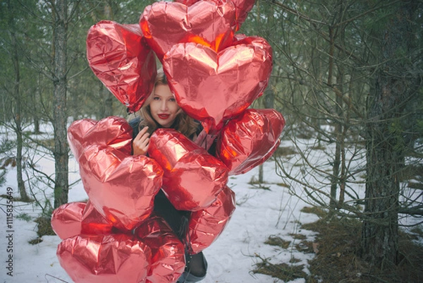 Fototapeta Romantic winter landscape with a woman holding a bunch of red balloons in the shape of hearts in a snowy forest among coniferous trees