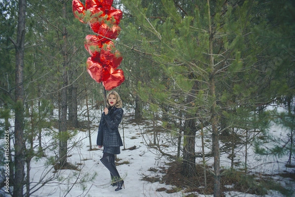 Fototapeta A woman standing in a winter forest holding a bunch of red balloons in the shape of hearts. There is snow on the ground and the trees are surrounded by green coniferous branches.