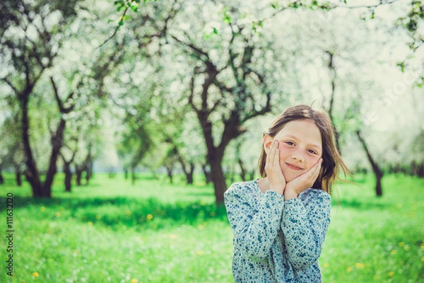 Fototapeta Smiling little girl in the garden, holding hands your face. Child in spring fruit orchard.