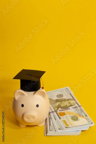 Fototapeta Vertical image of piggy bank with a mortarboard next to a wad of hundred dollar bills on an isolated yellow background, concept of saving for education