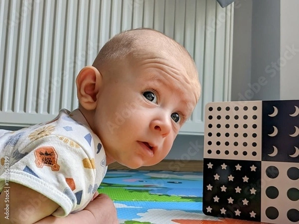 Fototapeta A 3-month-old baby lying on their tummy, attentively gazing at black-and-white contrast cards. The baby’s curious expression and focused gaze highlight early cognitive and visual development. 