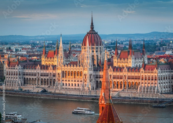 Fototapeta The photo captures the Hungarian Parliament Building in Budapest during sunset, beautifully reflecting the warm sunlight on its Neo-Gothic architecture along the Danube River.