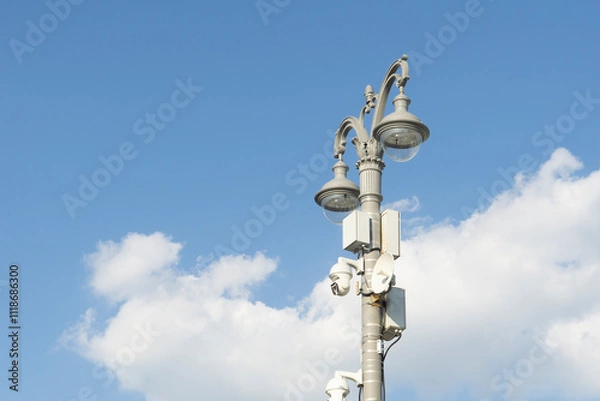 Fototapeta Surveillance cameras with repeater on a lamp post. Carved light pole against a cloudy sky.