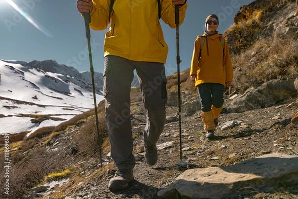 Fototapeta Two tourists in bright clothes walk against the background of snowy mountains in trekking shoes and trekking poles. Travelers in yellow clothes on the background of mountains go to the front.