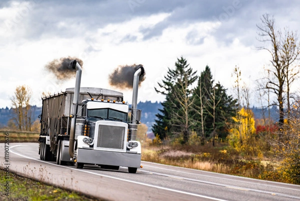 Obraz Black big rig classic semi truck with with thick black smoke coming from the exhaust pipes transporting cargo in bulk semi trailer running on the road polluting the environment