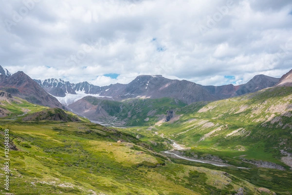 Fototapeta Wide green alpine valley with snake river among hills and rocks with vast view to large snow mountain top and big glacier tongue under cloudy sky. Serpentine river in high mountains. Shadows of clouds