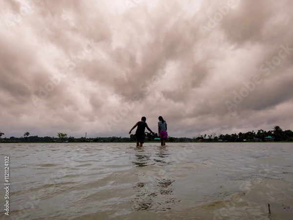 Obraz Silhouette of Two People Walking in Flooded Water Under Dramatic Cloudy Sky