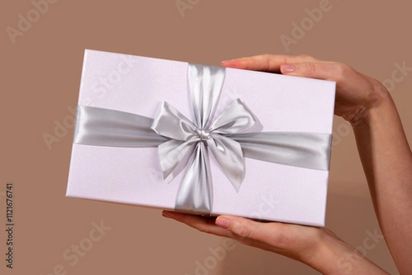 Fototapeta Womans hands holding a white gift box adorned with a silver ribbon bow against a rich brown background, symbolizing celebration, surprise, and special occasions Mocha Mousse color of 2025 year