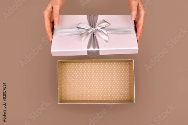 Fototapeta Female hands opening an empty gift box adorned with a silver ribbon bow set against a warm brown Mocha Mousse color of 2025 year background capturing the essence of surprise and celebration from above