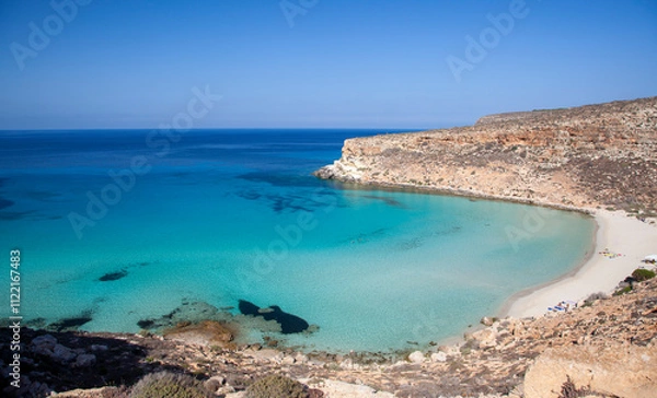 Fototapeta A breathtaking view of Spiaggia dei Conigli in Lampedusa, featuring crystal-clear turquoise waters, white sand, and rugged cliffs under a bright blue sky.