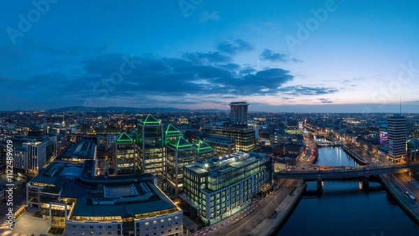 Fototapeta A dusk view of Dublin's modern skyline with the iconic green-lit Bank of Ireland building, a river reflecting the city lights, and a bridge crossing over it.