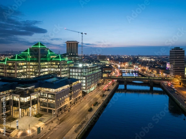 Fototapeta A dusk view of Dublin's modern skyline with the iconic green-lit Bank of Ireland building, a river reflecting the city lights, and a bridge crossing over it.