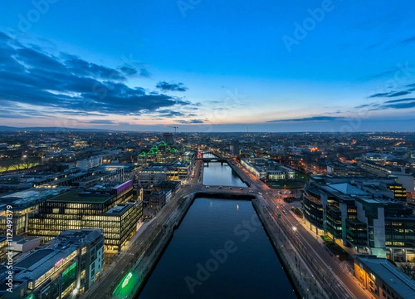 Fototapeta A dusk view of Dublin's modern skyline with the iconic green-lit Bank of Ireland building, a river reflecting the city lights, and a bridge crossing over it.