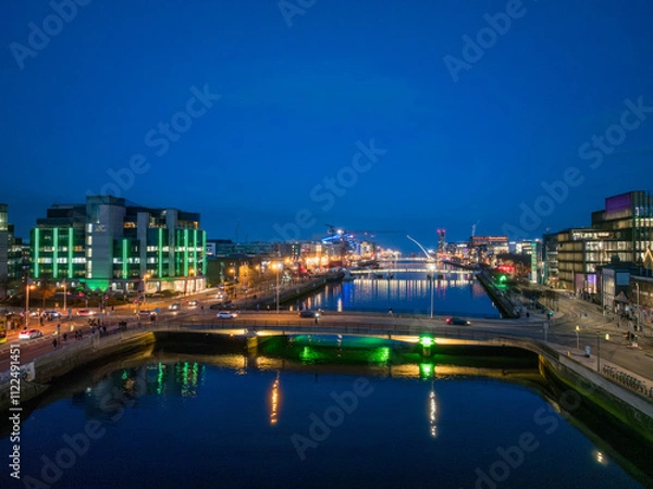 Fototapeta A dusk view of Dublin's modern skyline with the iconic green-lit Bank of Ireland building, a river reflecting the city lights, and a bridge crossing over it.