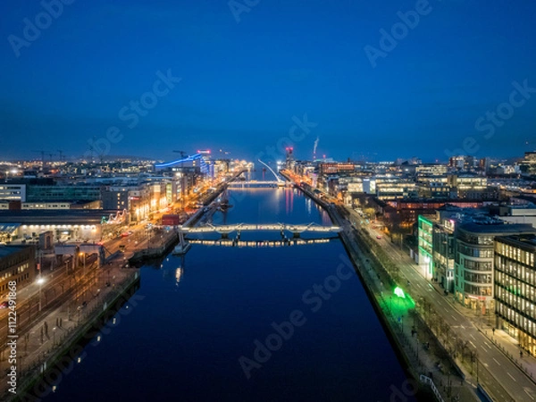 Fototapeta A dusk view of Dublin's modern skyline with the iconic green-lit Bank of Ireland building, a river reflecting the city lights, and a bridge crossing over it.