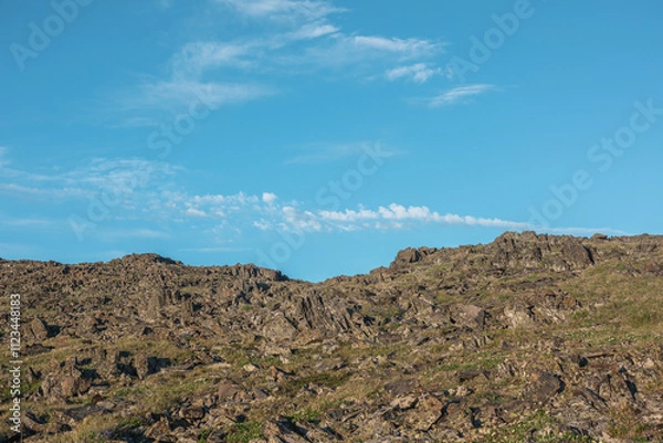Fototapeta Nature layered background with sunlit grassy rocky hill under clouds in blue sky. Minimalist view to stone hill in bright sun in changeable weather. Natural backdrop of rock mountain pass in sunny day