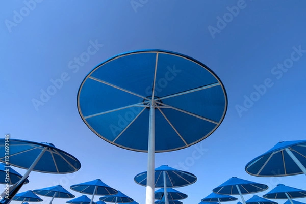 Fototapeta Blue parasols from below on the beach and pool under a clear blue sky with white painted poles at holiday location