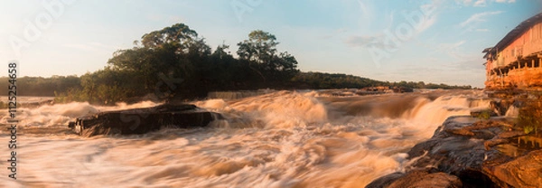 Obraz Rushing waters of the Guaviare River create powerful cascades amidst dense greenery as twilight descends upon the Amazon rainforest, showcasing nature's raw beauty and splendor.