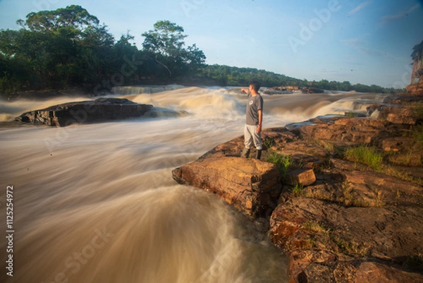 Obraz Standing on rocky terrain, an individual gazes at the powerful currents of the Guaviare River. Bustling waters reflect the soft glow of the early morning sun against lush greenery