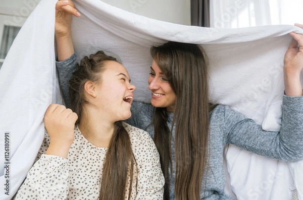 Obraz A young woman with Down syndrome and her mother are sharing a playful moment under a white sheet. They are laughing and looking at each other, showcasing a loving and supportive relationship. 