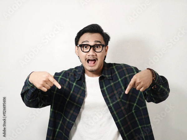 Obraz Smiling young handsome Asian man wearing a plaid shirt pointing fingers down, showing free place for advertisement isolated on white background. People lifestyle concept