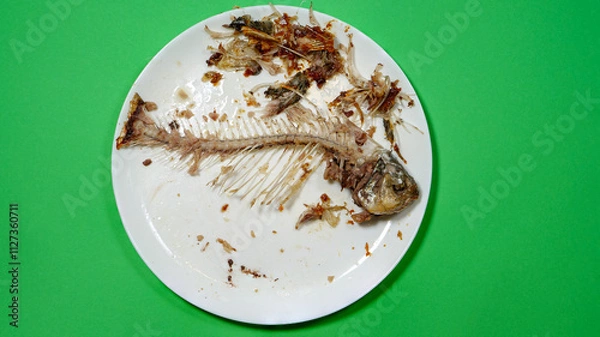 Fototapeta Fish Bones on The White Plate and Green Background. The Food Waste. Remains of Fried Fish. Fishbone and Leftover Meat of Crucian Carp After Eat