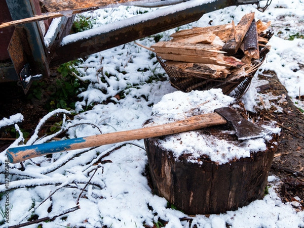 Fototapeta The axe lies on a wooden stump with a pile of chopped firewood on the snow-covered ground. Heating the house with wood.