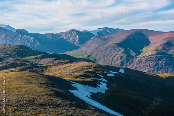 Fototapeta Scenic sunny aerial top view to sunlit big green hills against bright high rocky mountain range in bright sun under clouds in blue sky. Light and shadows on colorful large mountains just before sunset
