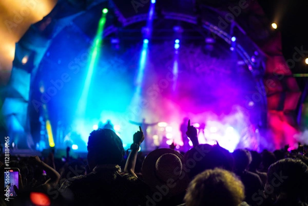 Obraz Silhouettes of crowd at concert in front of stage with bright spotlights.The back view of people enjoy live music at the band's performance. Outdoor concert with musicians on stage and fans.